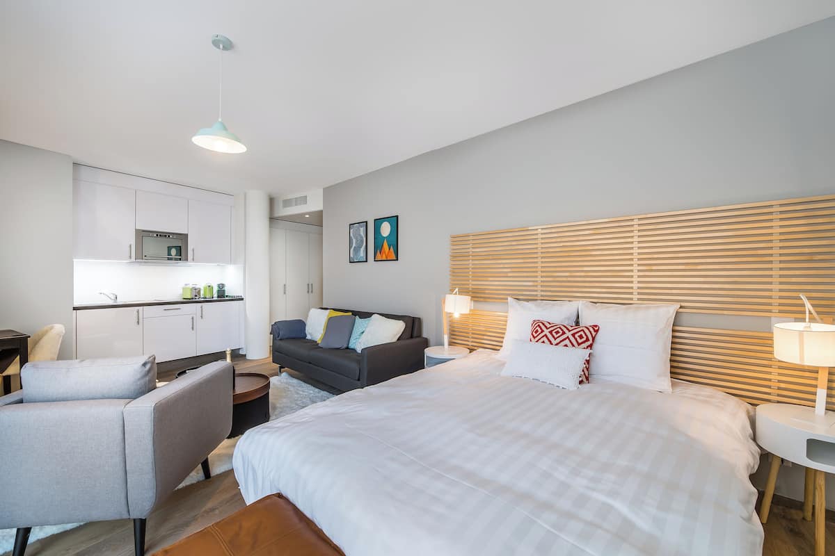 A modern hotel room with a large bed, a sofa, a small kitchen area, and a dining table.