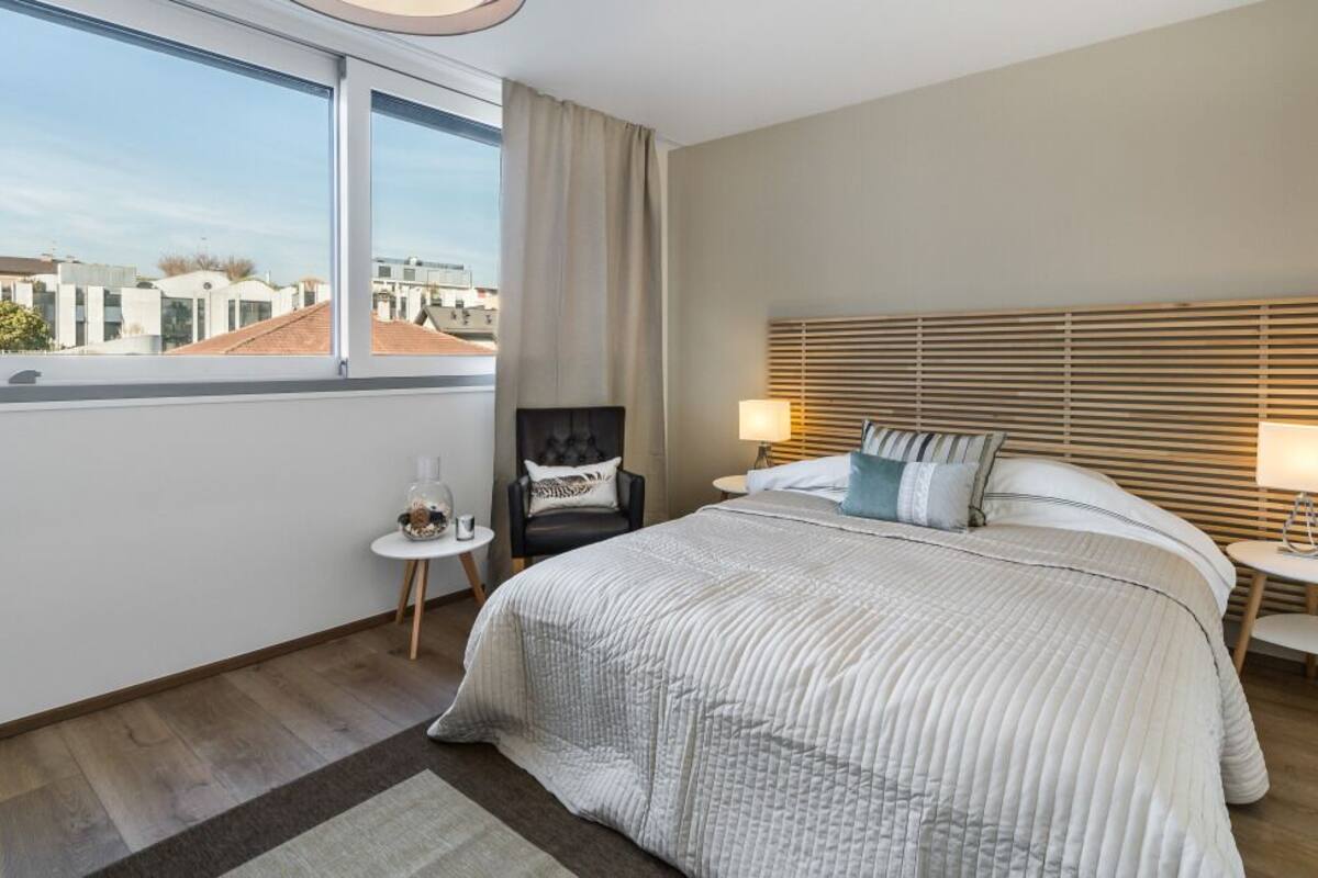 A modern hotel room with a large bed, two bedside tables, a chair, and a view of the city through large windows.