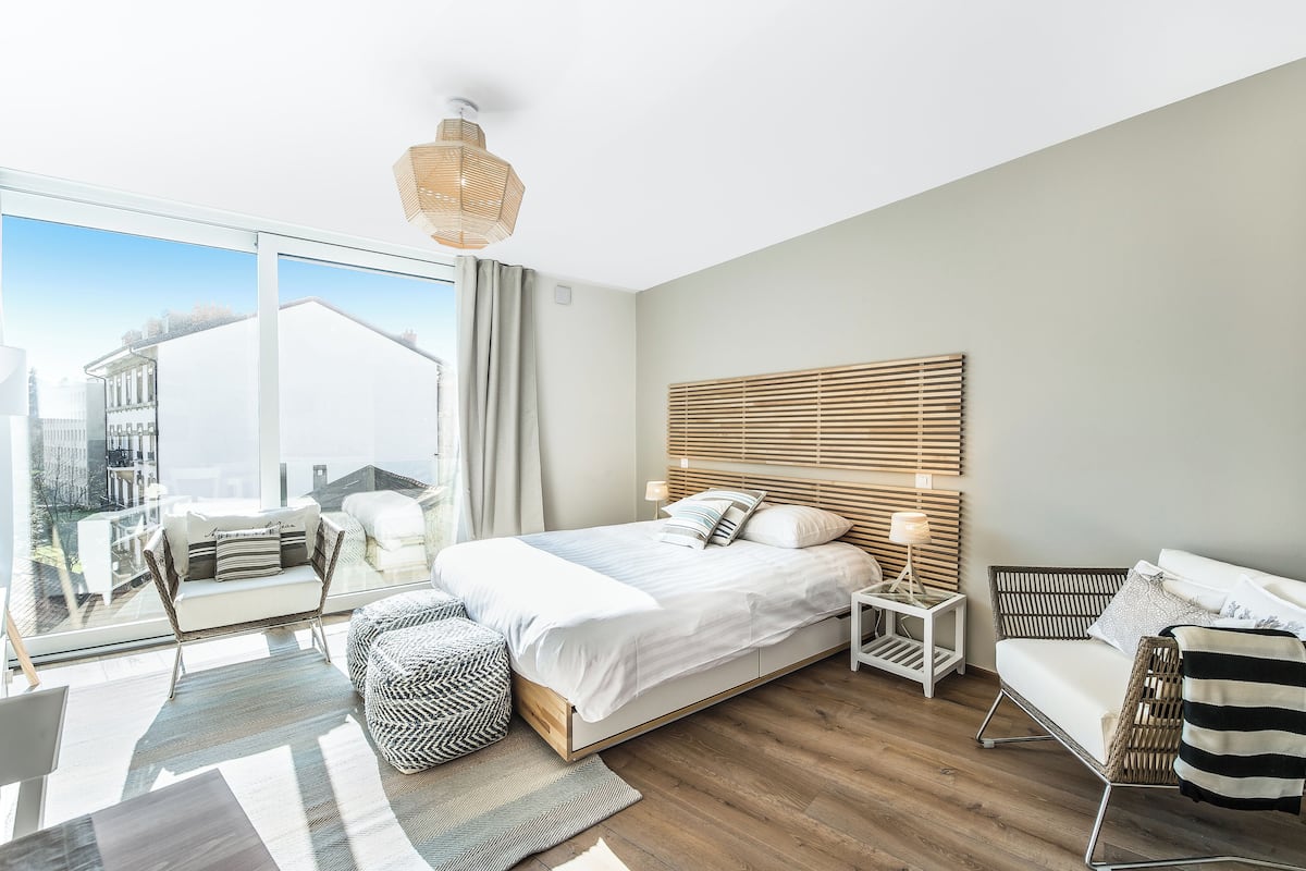 A modern hotel room with a large bed, a seating area with a sofa and armchair, a small side table, and a view of the city through large windows.