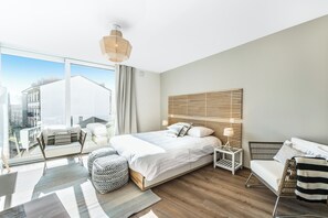 Une chambre d’hôtel moderne dotée d’un grand lit, d’un coin salon avec un canapé et un fauteuil, d’une petite table d’appoint et offrant une vue sur la ville grâce à de grandes fenêtres.