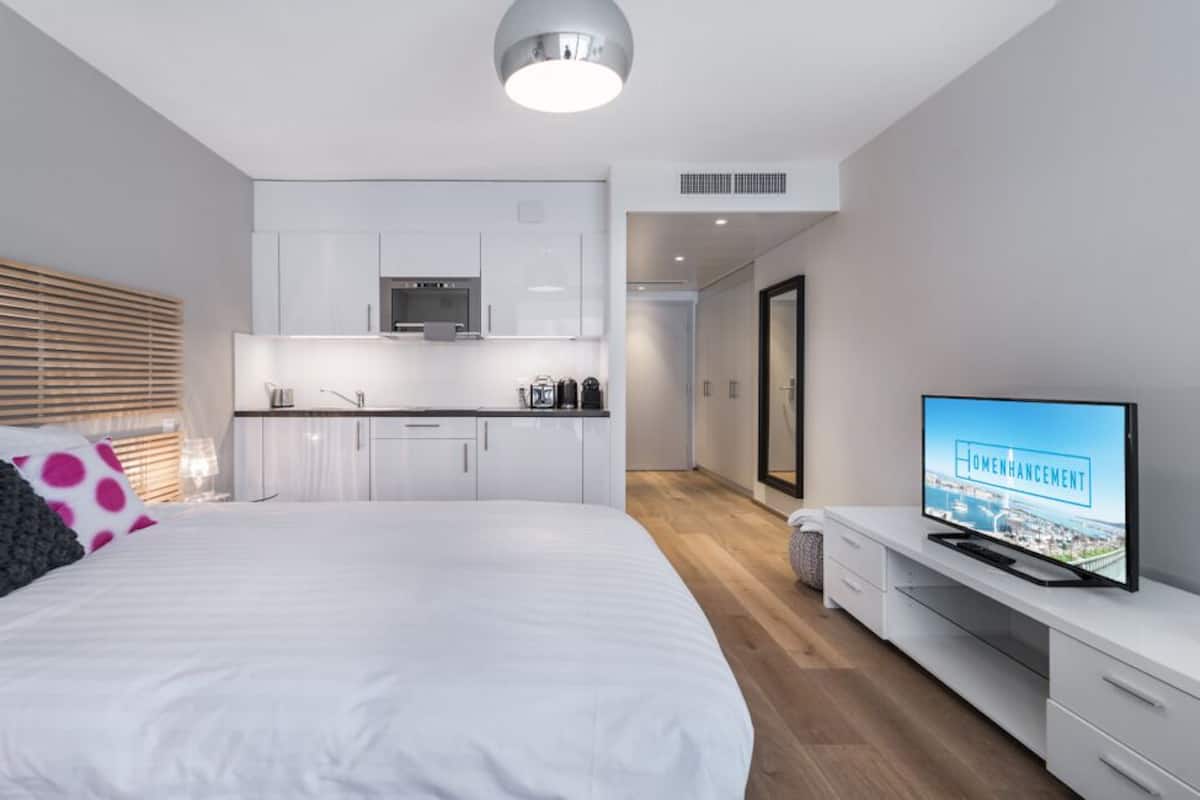 A modern hotel room with a large bed, a TV, a kitchenette, and a bathroom.