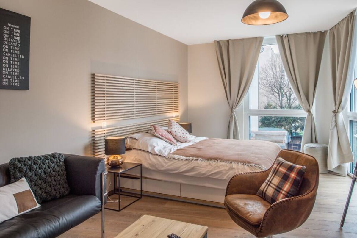 A bedroom with a bed, a nightstand, a lamp, a chair, and a window with curtains.