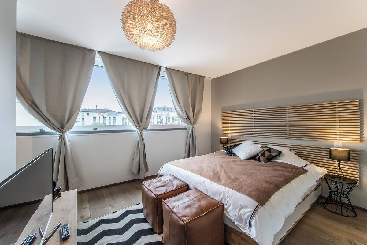 A modern bedroom with a large bed, a desk with a computer, and a view of buildings through the window.