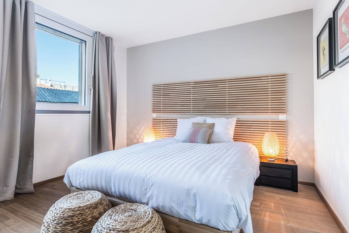 A bedroom with a large bed, a nightstand with a lamp, a window with curtains, and a view of buildings outside.