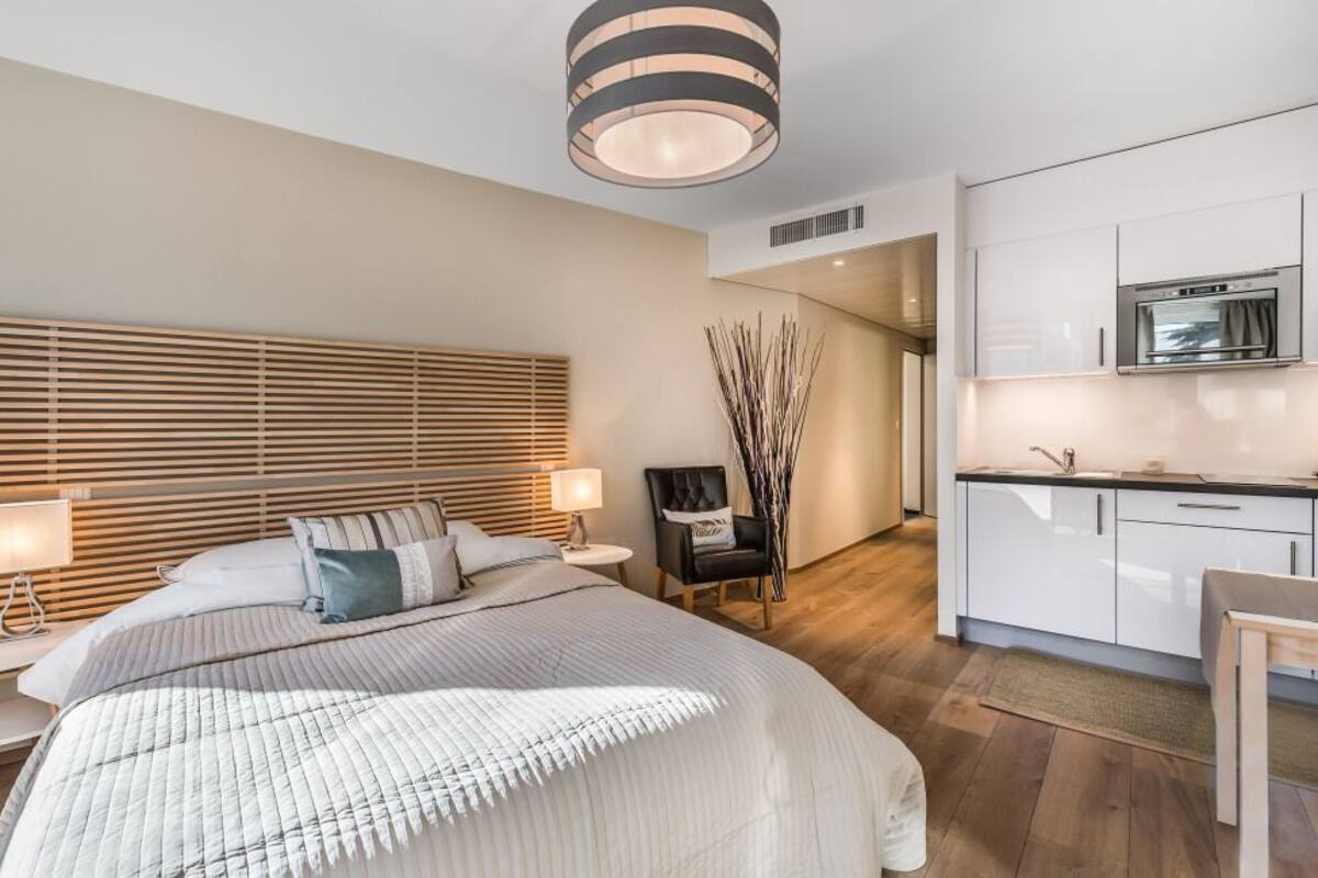 A modern bedroom with a large bed, bedside tables, a desk, and a kitchenette.