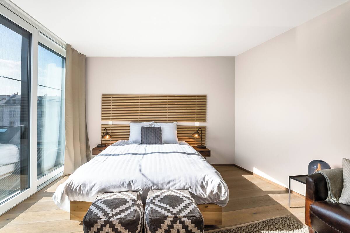 A modern bedroom with a large bed, a bedside table with a lamp, a window with curtains, and a view of the city.