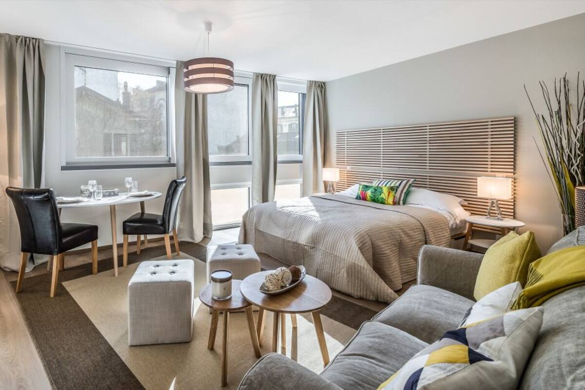 A modern hotel room with a bed, a sofa, a dining table with chairs, a TV, and a large window with curtains.