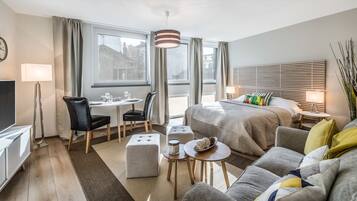 A modern hotel room with a bed, a sofa, a dining table with chairs, a TV, and a large window with curtains.