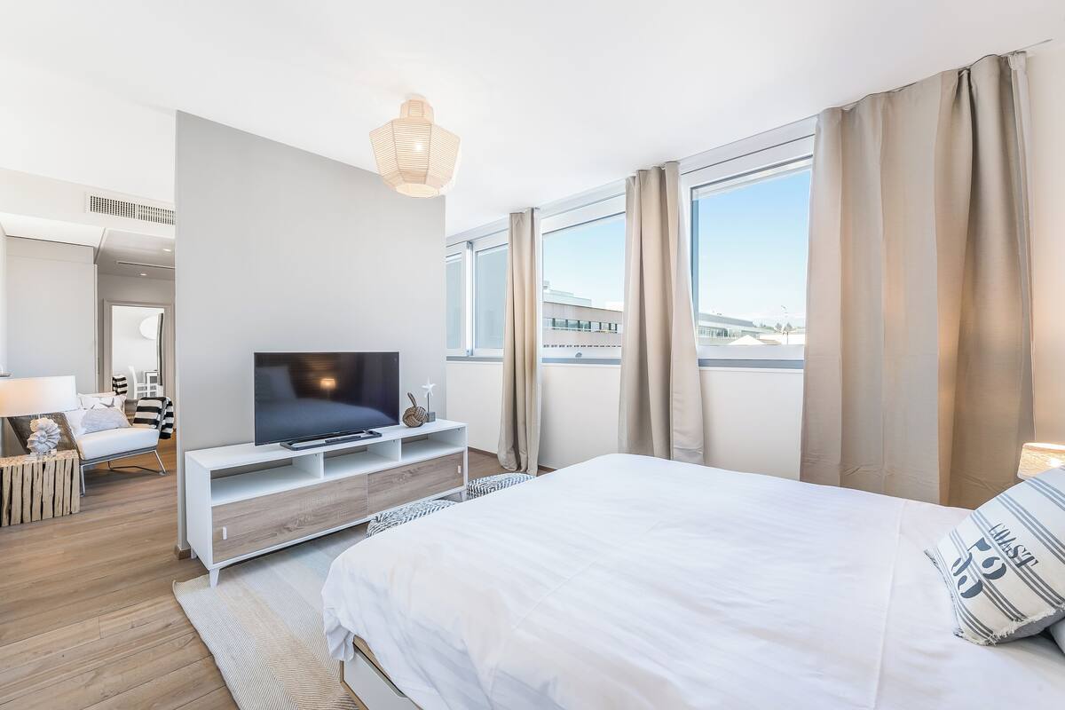 A modern hotel room with a large bed, a TV on a white console, a view of the city through large windows, and a small seating area with a lamp and decorative items.