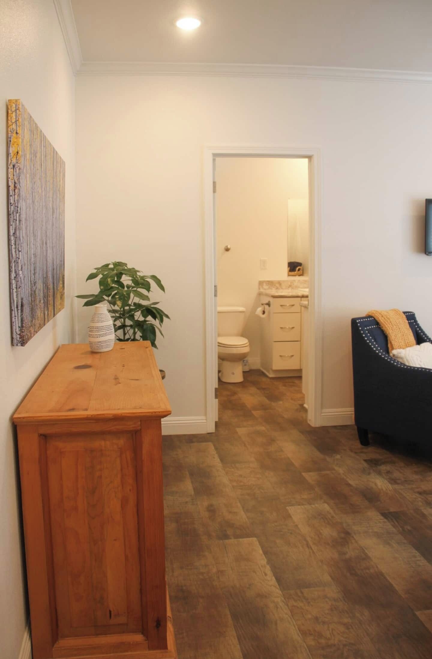 A hallway leading to a bathroom with a wooden floor, a small table with a vase and plant, and a painting on the wall.