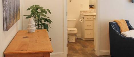 A hallway leading to a bathroom with a wooden floor, a small table with a vase and plant, and a painting on the wall.