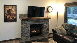 A living room with a stone fireplace, a flat-screen TV mounted above it, a clock on the wall, and a window with blinds.