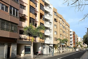 A multi-story residential building with balconies, a ground-level commercial space, and palm trees.