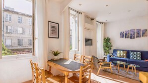 Una sala de estar luminosa con comedor, ventanas amplias y vista a edificios.