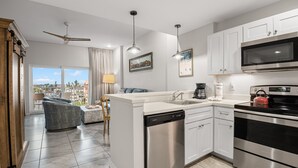 A modern kitchen with white cabinets, stainless steel appliances, and a dishwasher. There is a dining area with a sofa and a view of the city.