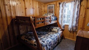A bunk bed with a patterned bedspread, wooden walls, a window with curtains, and a painting on the wall.
