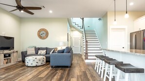 Un salon moderne avec un canapé, un pouf et un téléviseur. Un escalier mène à une cuisine équipée d’un bar avec des tabourets.