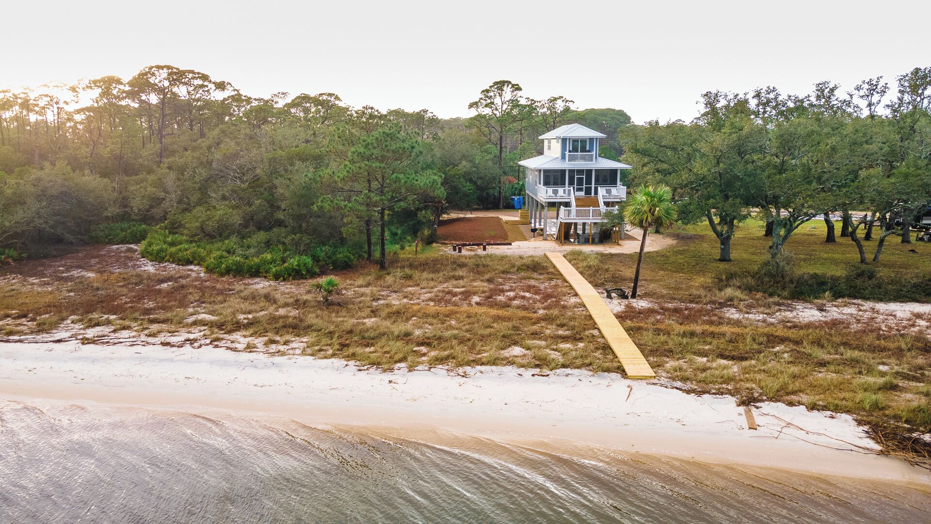 Une maison à deux étages avec un balcon, entourée d’arbres et bordée d’une plage de sable.