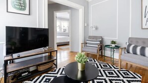 A modern living room with a flat-screen TV, a coffee table, a sofa, and a chair. There is a framed quote on the wall and a door leading to another room.