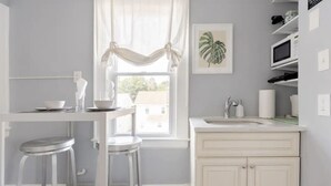 A kitchen with a white countertop, a sink, and a window with curtains. There are two stools and a framed plant on the wall.