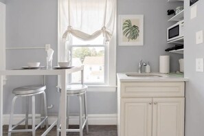 A kitchen with a white countertop, a sink, and a window with curtains. There are two stools and a framed plant on the wall.
