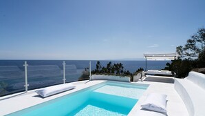 Ein Pool mit Blick auf das Meer, klares, blaues Wasser, weiße Liegestühle und eine Pergola.