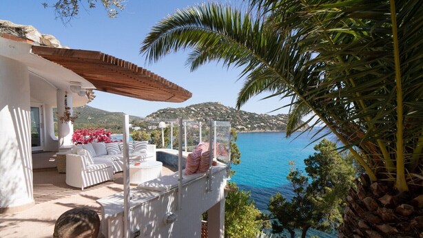 Eine Terrasse mit weißer Möbel, Blick auf das Meer und eine gebirgige Landschaft.
