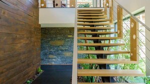 A modern staircase with wooden steps and metal railings, leading to a second floor with a stone wall and a view of greenery outside.
