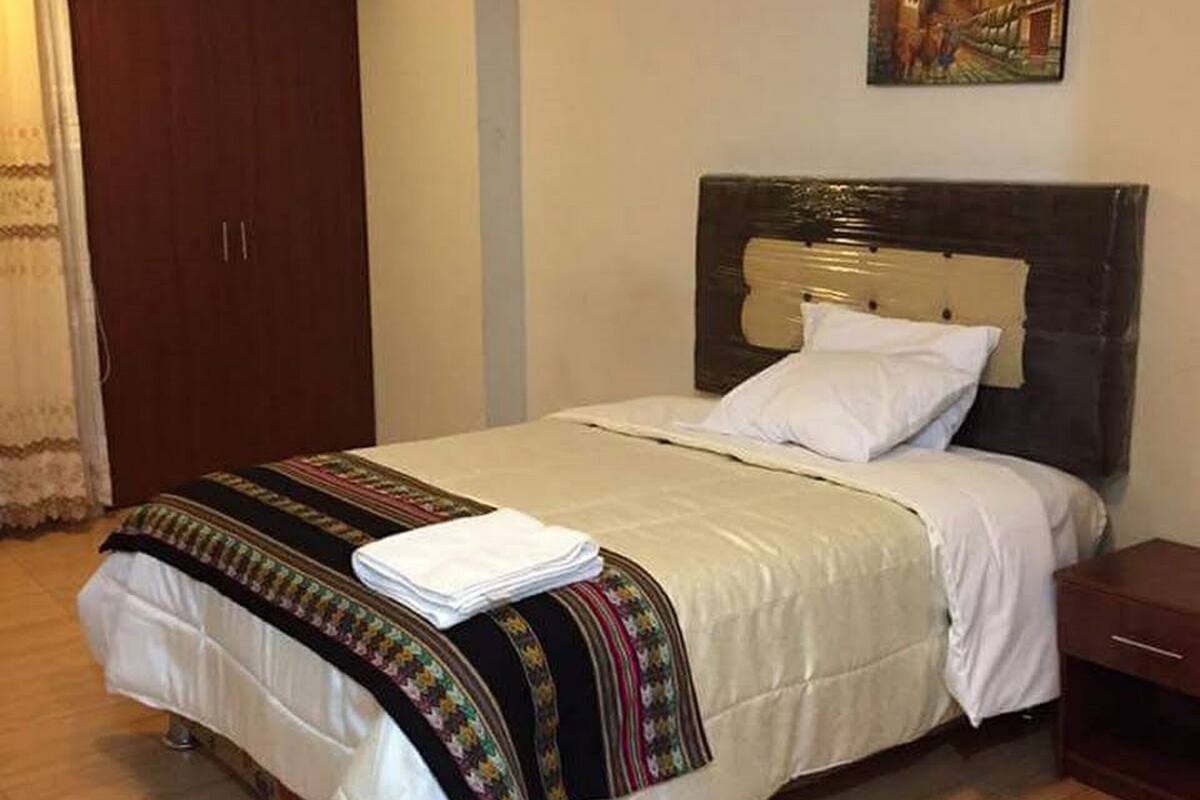 A neatly made bed with a white comforter and a colorful blanket, a wooden headboard, a small bedside table, and a painting on the wall.