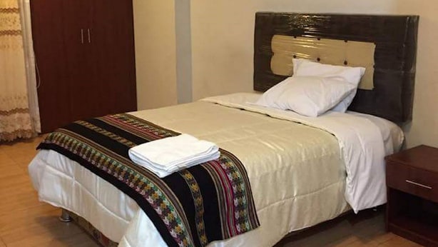 Una cama bien hecha con edredón blanco y manta colorida, cabecera de madera, mesita de noche y un cuadro en la pared.