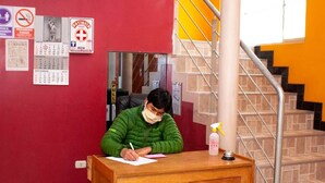 Una persona sentada en un escritorio de madera en una habitación con pared roja, una escalera y varios carteles, incluyendo un letrero de prohibido fumar.