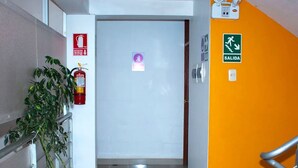 Un pasillo con extintor, planta en maceta, puerta con letrero y escalera con barandilla.