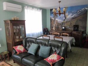 Ein Wohnzimmer mit Ledersofa, Esstisch und Stühlen. Es gibt ein Sideboard mit Glastüren und ein Bild an der Wand.