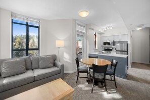 A modern kitchen with a central island, a dining area with a round table and chairs, a grey sofa, and a large window offering a view of trees.