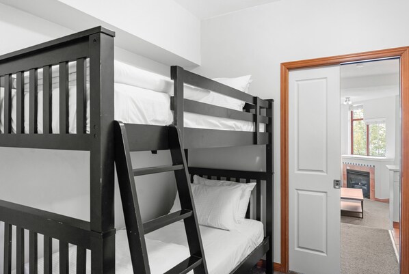 A bunk bed with white bedding, a wooden ladder, and a door leading to a room with a fireplace.