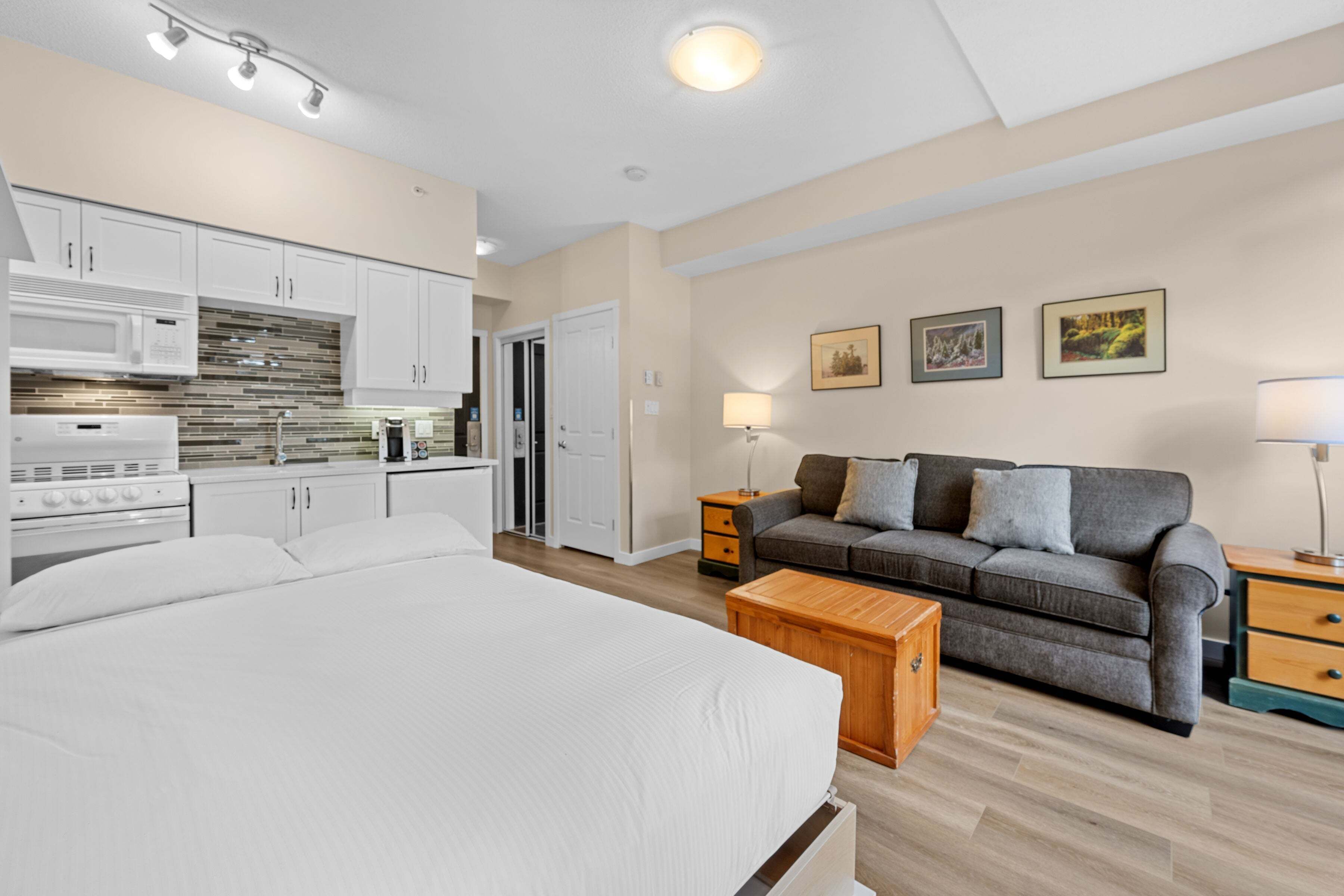 A bedroom with a large bed, a sofa, a wooden nightstand, and a kitchen area with cabinets and appliances.