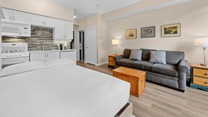 Een slaapkamer met een groot bed, een slaapbank, een houten nachtkastje en een keukenhoek met kastjes en apparatuur.