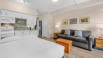 A bedroom with a large bed, a sofa, a wooden nightstand, and a kitchen area with cabinets and appliances.