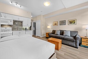 Un dormitorio con una cama grande, un sofá, una mesita de noche de madera y una zona de cocina con armarios y electrodomésticos.