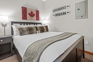A neatly made bed with a white comforter and two pillows, a wall-mounted light, a framed Canadian flag, and a sign that reads 'WHISTLER EXPERT'.