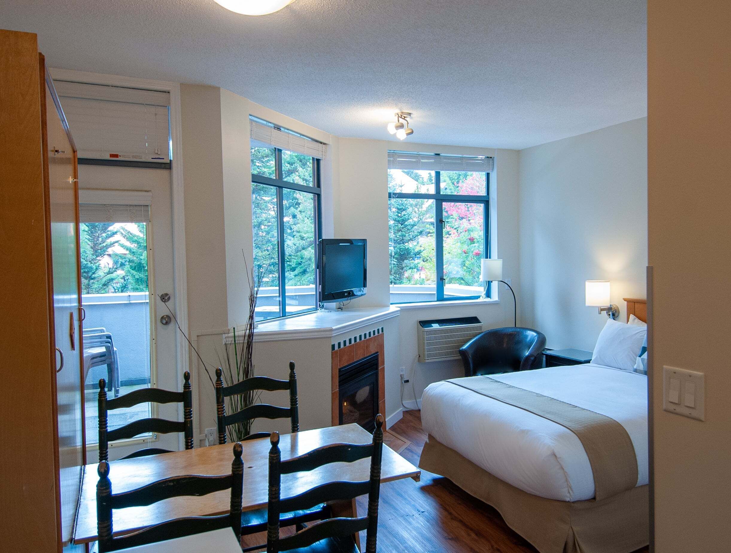 A bedroom with a bed, a desk with a computer, a chair, and a TV. There are windows with blinds, a fireplace, and a door.