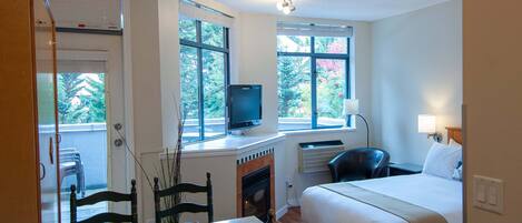 A bedroom with a bed, a desk with a computer, a chair, and a TV. There are windows with blinds, a fireplace, and a door.