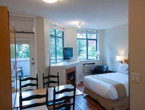 A bedroom with a bed, a desk with a computer, a chair, and a TV. There are windows with blinds, a fireplace, and a door. - Alpenglow Lodge Legacy Studio by MVA (Whistler)