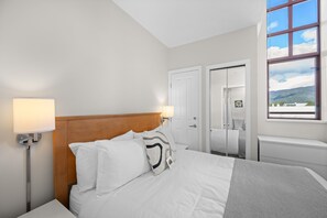 A hotel room with a bed, bedside lamps, a window with a view, and a door leading to another room. - Alpenglow Lodge Junior Penthouse by MVA (Whistler)