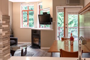 A modern kitchen with a fireplace, a dining table with chairs, a television mounted on the wall, and a view of greenery outside.