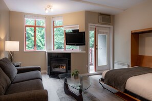 A modern hotel room with a fireplace, a flat-screen TV, a sofa, a bed, and large windows with a view of greenery.