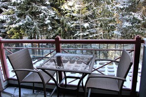 A balcony with two wicker chairs and a glass table, overlooking snow-covered trees.