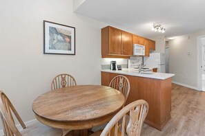 A kitchen with wooden cabinets, a white countertop, and a dining area with a wooden table and chairs. - Alpenglow Lodge Bear Wonder One-Bedroom by MVA (Whistler)