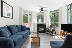 Ein Wohnzimmer mit einer Couch, einem Glastisch, einem Kamin, einem Fernseher und großen Fenstern.