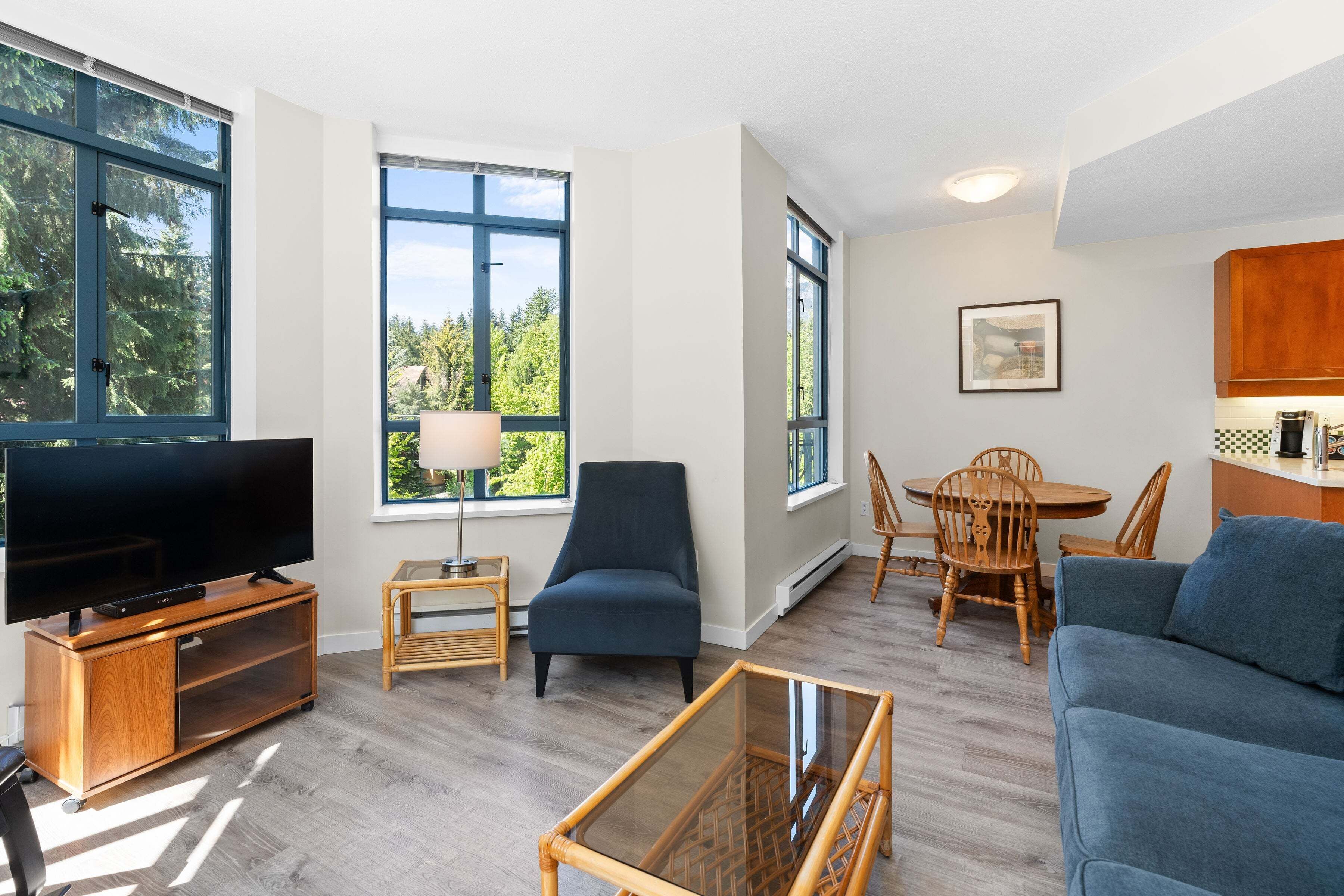 Een woonkamer met een televisie, een blauwe bank, een glazen salontafel, een donkere fauteuil, een eettafel met stoelen en grote ramen met uitzicht op bomen.